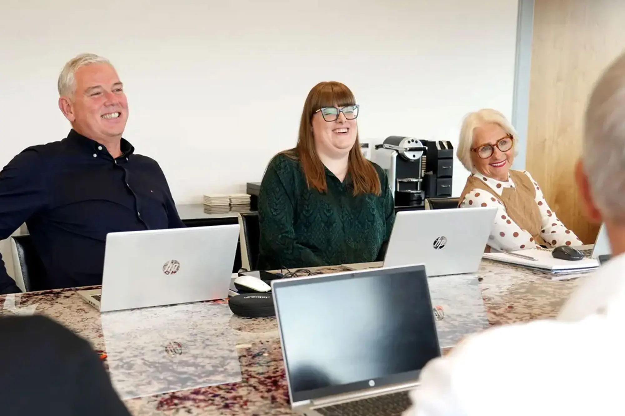 Three people sat at a meeting table with laptops, smiling mid conversation
