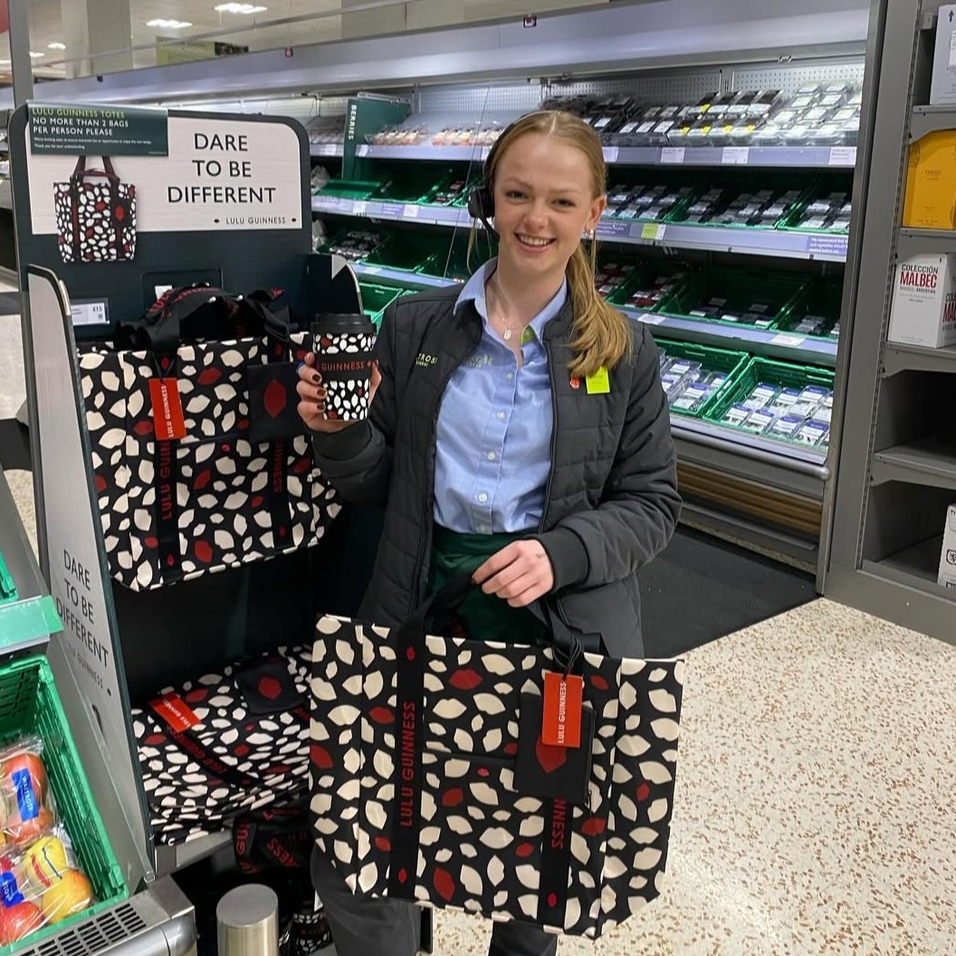 A supermarket worker wearing uniform stood in an aisle holding a branded bag and cup, smiling at the camera. .