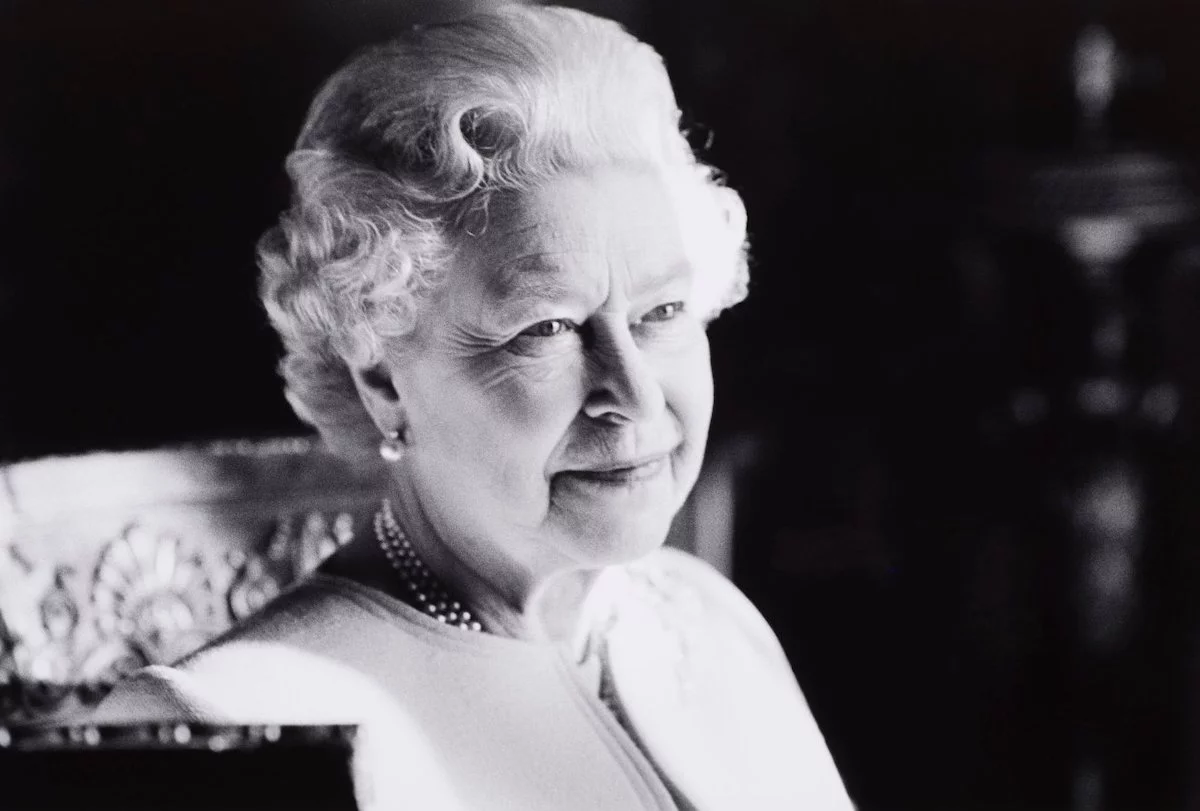 A black and white headshot of Her Majesty The Queen smiling. 